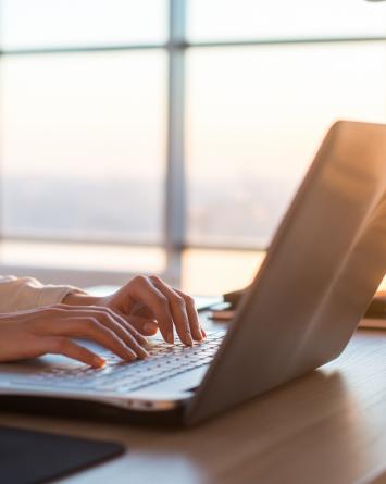 A businesswoman types on a laptop as the sun sets through the large windows behind her