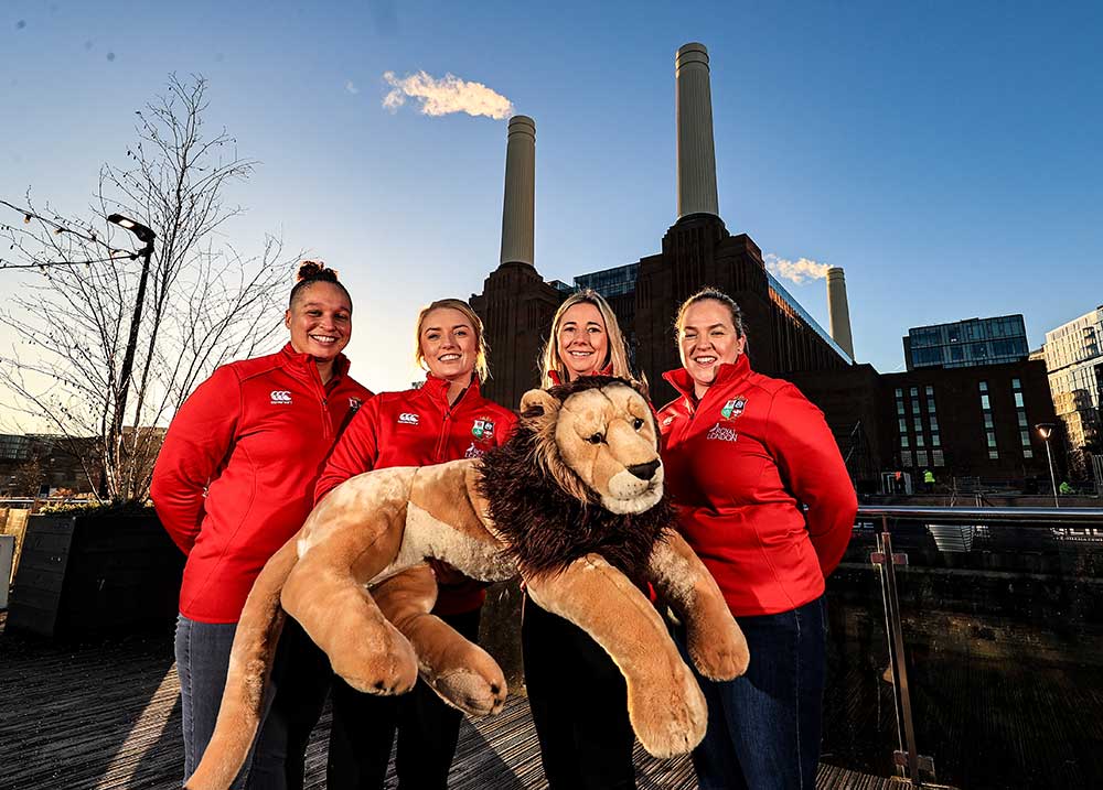 Four women in red jackets stand outside with a large plush lion, with an industrial building in the background.