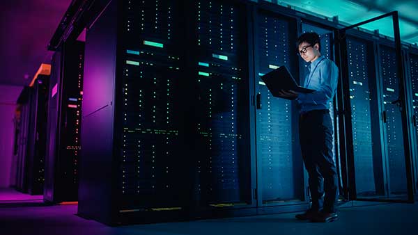 A person stands in a server room holding an open laptop, surrounded by illuminated server racks.
