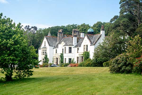 A large white manor house surrounded by trees and a green lawn under a partly cloudy sky.
