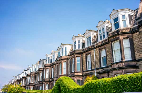 Row of Victorian-style terraced houses with bay windows under a clear blue sky and green hedges in front.