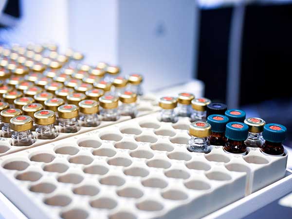 Rows of small glass vials with colored caps are arranged in white trays with circular slots on a laboratory bench.