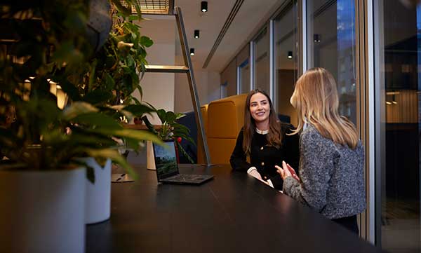 Two women sit and talk at a table in a modern office space, with plants and a laptop visible in the foreground.