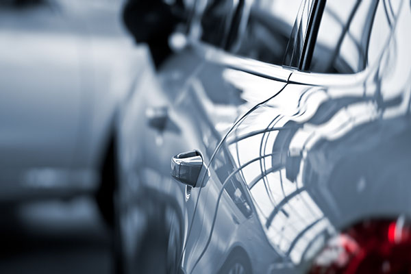 Close-up of a shiny car door and handle with building reflections, with another blurred car in the background.