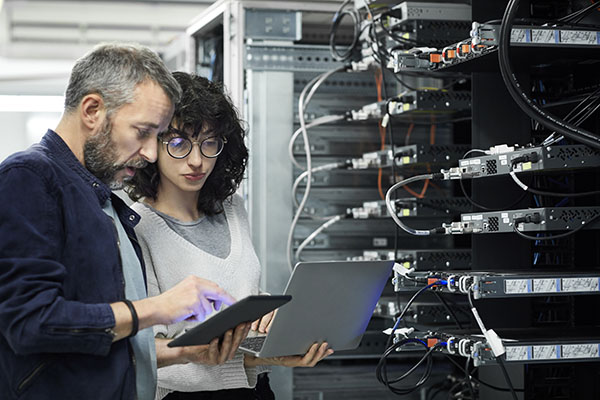 Two people check a tablet and laptop while standing next to server racks in a data center or server room.