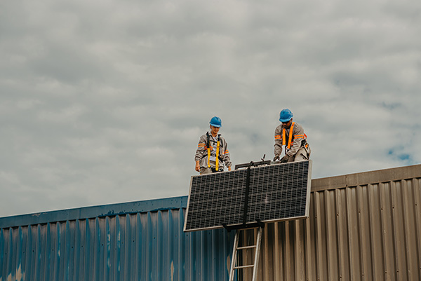 Two workers in safety gear install a solar panel on the roof of a building under a cloudy sky.