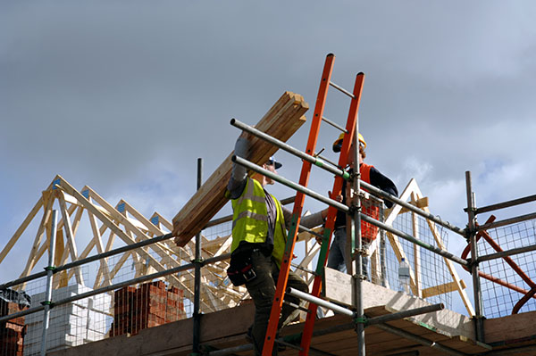 Two construction workers in safety gear carry wooden beams on scaffolding at a building site with roof trusses in the background.
