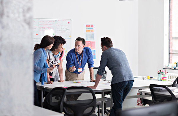Four people stand around a table in an office, discussing documents and notes on a whiteboard.