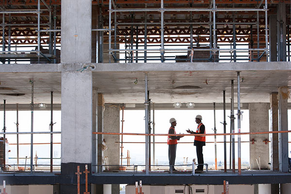 Two construction workers wearing helmets and vests talk on the second floor of a partially built concrete structure.