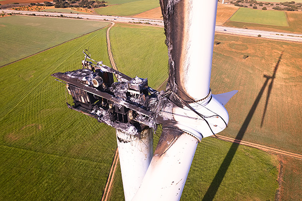 A wind turbine with a severely burned and damaged nacelle stands above green and brown farmland.