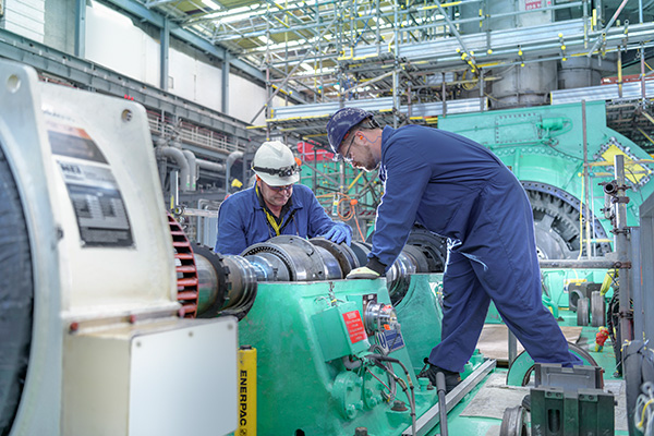 Two engineers in safety gear inspect and work on large industrial machinery inside a factory setting.