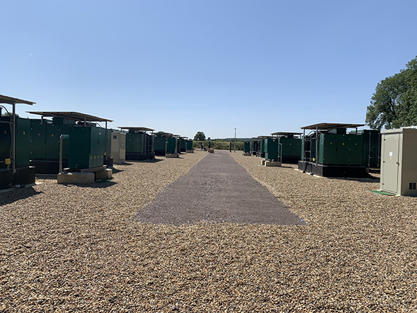 A row of large green electrical generators on gravel, separated by a wide central pathway under a clear sky.
