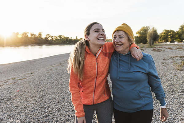 Two women in athletic wear smiling and walking arm in arm on a rocky riverside path at sunset.
