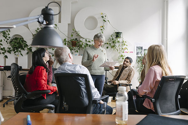 Five people in an office sit in a casual meeting, one woman standing and speaking while others listen attentively.