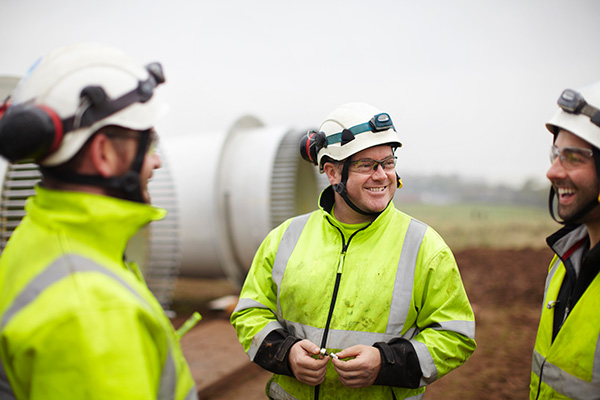 Three workers in high-visibility jackets and helmets converse outdoors at an industrial or construction site.