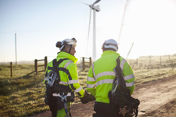 Two workers in high-visibility gear and safety harnesses stand outdoors near a wind turbine on a dirt path.