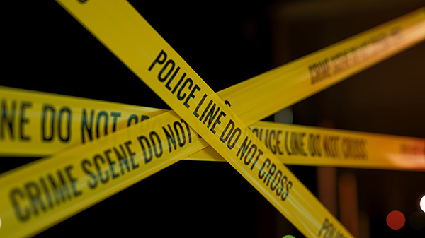 Yellow police tape reading "Do Not Cross" blocks off a crime scene area with a dark background.