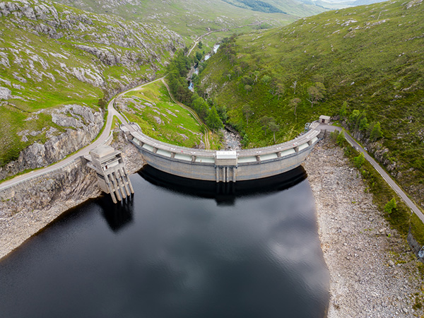 Aerial view of a concrete dam holding back a reservoir, surrounded by rocky and grassy hills.