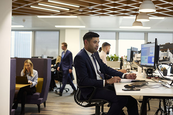 Man in a suit works at a computer in a modern office; colleagues are working at desks and in booths in the background.