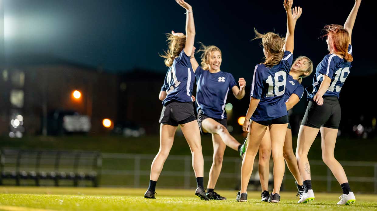 Women in blue jerseys celebrate on a football pitch under stadium lights at night.