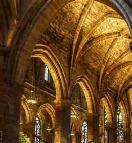 Arched stone ceiling and pillars inside a historic cathedral, with warm lighting and stained glass windows visible in the background.