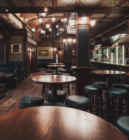 Empty pub interior with wooden tables, green leather stools, booth seating, and a long bar with taps and mirrors.