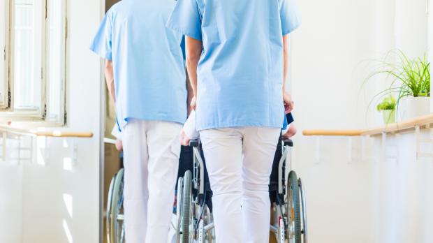Two care workers in nurse scrubs push two wheelchairs through the corridor of a care home
