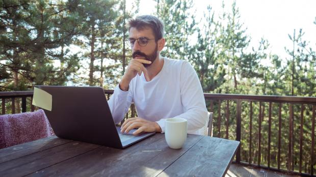 Man on balcony in the woods, working at laptop on wooden table