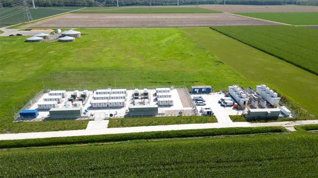 Aerial view of a green field with a white industrial facility, wind turbines in the background, and vehicles parked around the site.