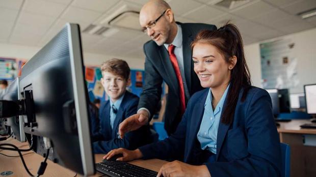 Teacher assisting two students working at computers in a classroom, all wearing formal school uniforms.