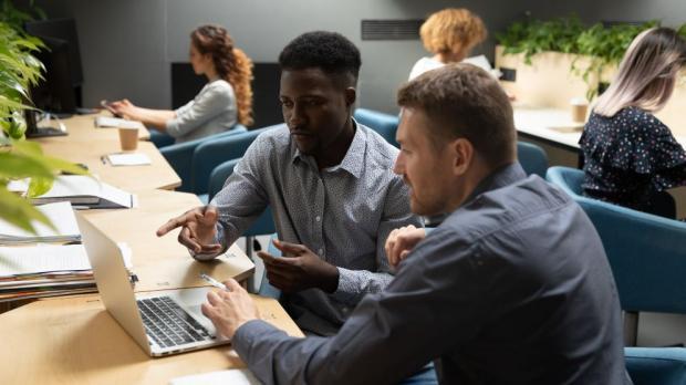 Focused african and caucasian partners colleagues mentor intern discuss online project using laptop in modern office, male diverse executive team two businessmen work in teamwork looking at computer