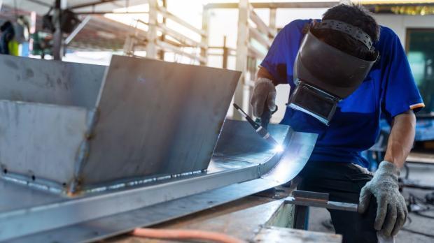 Industrial worker using TIG welding equipment on a curved metal structure in a factory. The welder wears safety gloves and a protective helmet in a workshop setting.