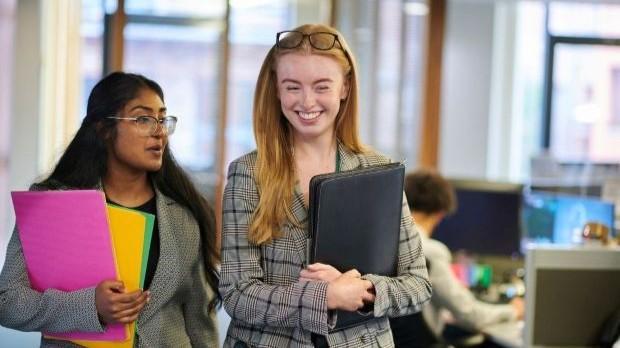 Two women in business attire holding folders walk through an office, one smiling and the other talking.
