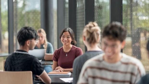 A group of students sit together at a table in a bright room, with one student smiling at the camera.