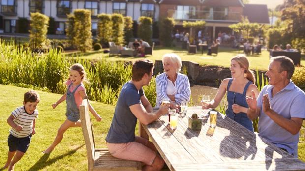 Three adults and two children sit at an outdoor table, while two kids run on grass in a sunny garden near a building.
