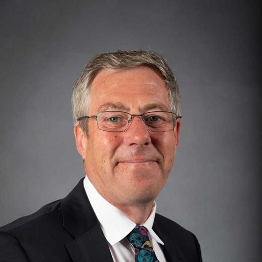 A middle-aged man with glasses wearing a dark suit, white shirt, and a colourful patterned tie, posing against a plain grey background.