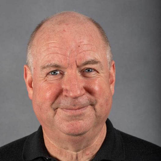 Bald man with a black polo shirt smiling slightly, standing in front of a plain gray background.
