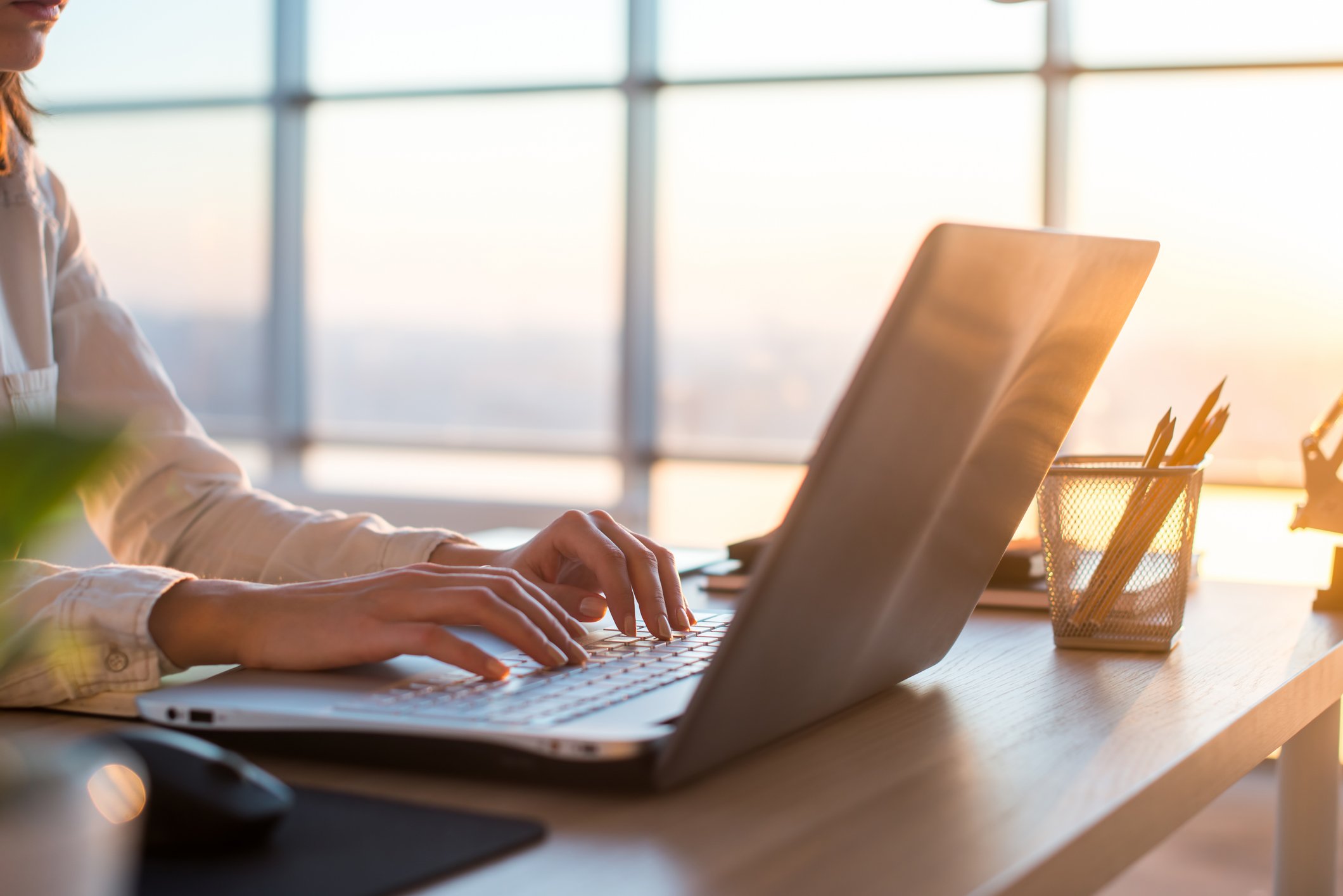 A businesswoman types on a laptop as the sun sets through the large windows behind her
