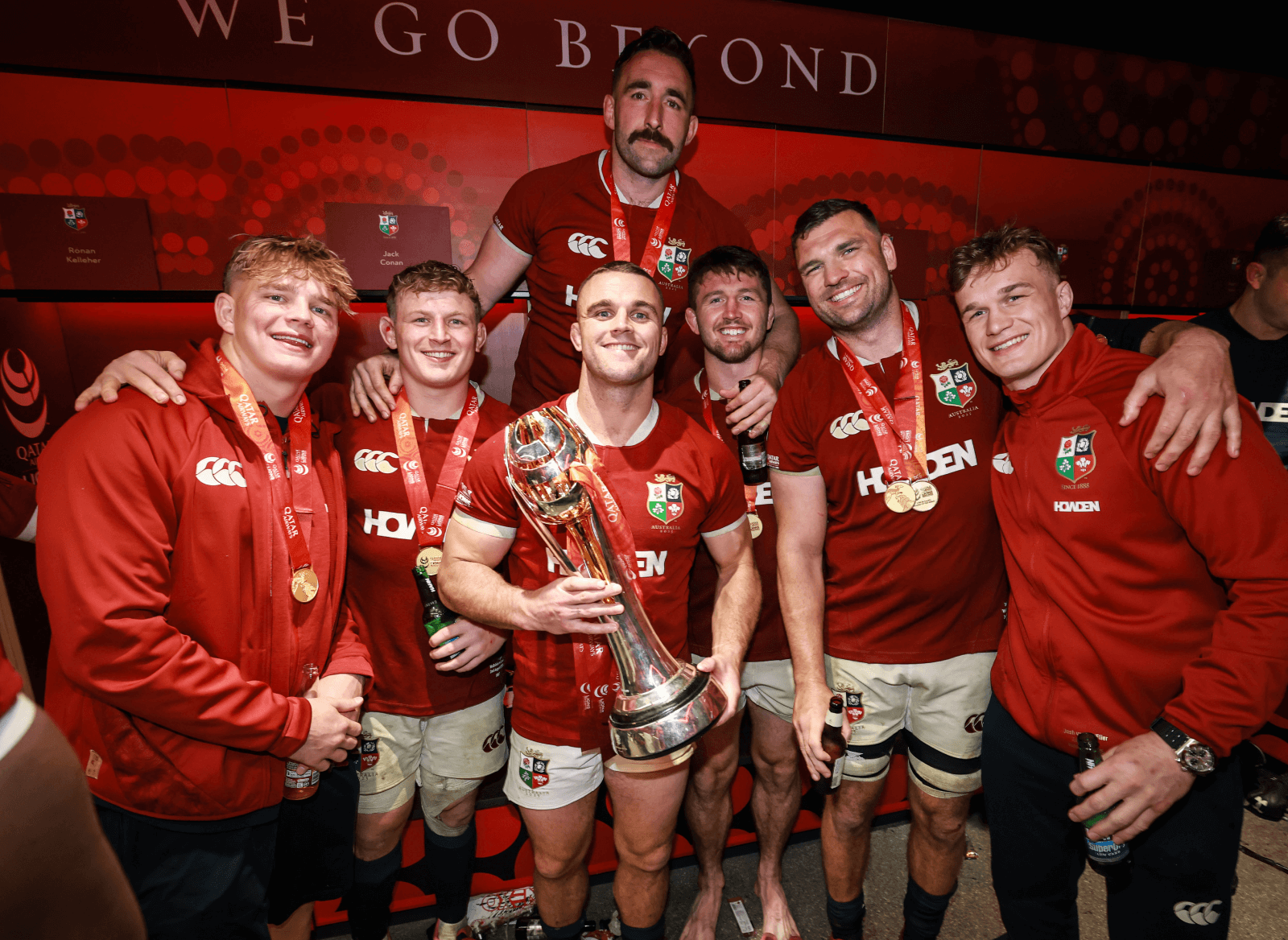 Seven rugby players in red jerseys pose with medals and a large trophy, smiling for a group photo in front of a red background that reads "WE GO BEYOND".