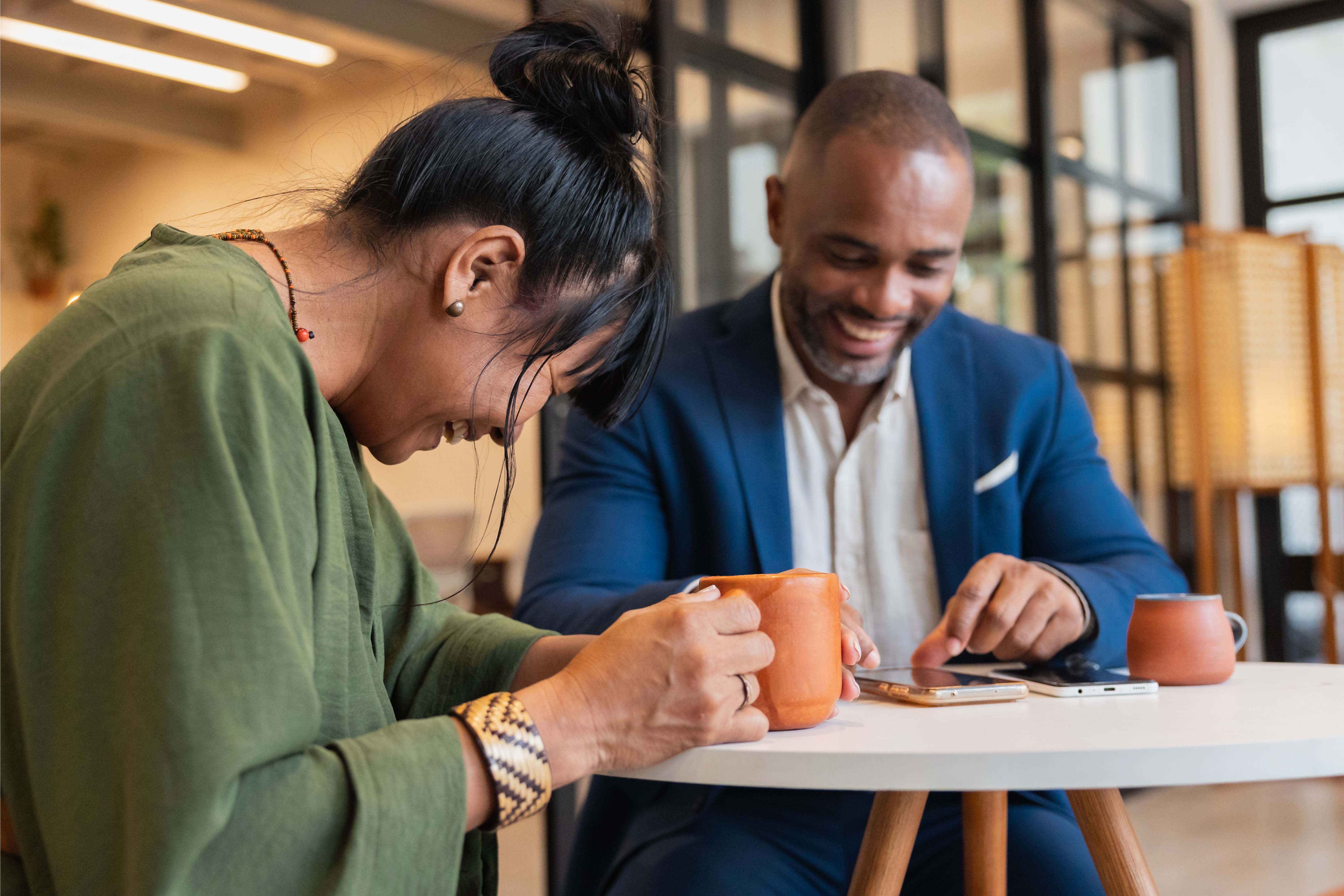 Man and woman smiling and laughing while having coffee and looking at a phone during a relaxed office catch-up