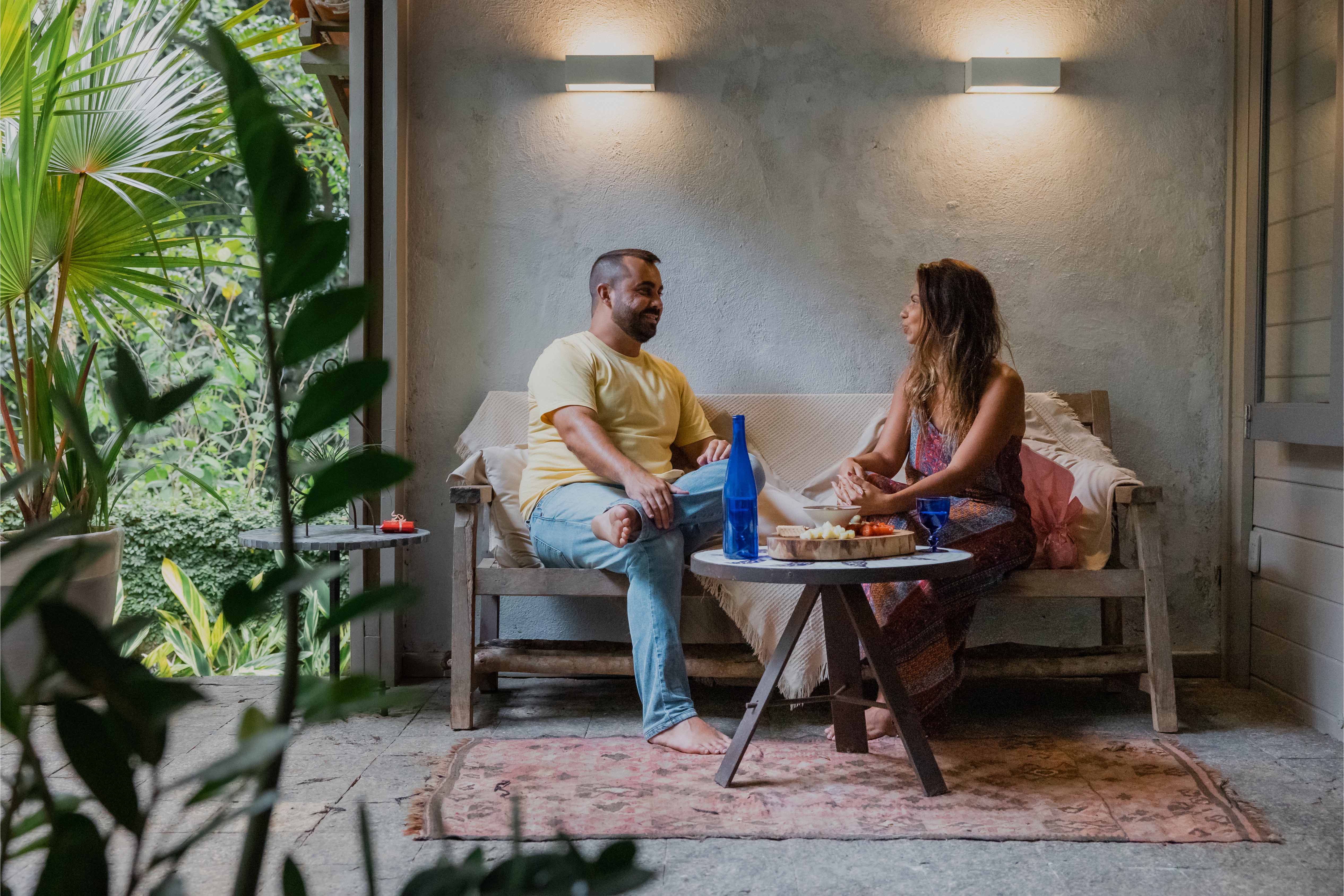 Man and woman sitting on a bench outdoors, sharing food and conversation in a cosy, plant-filled veranda