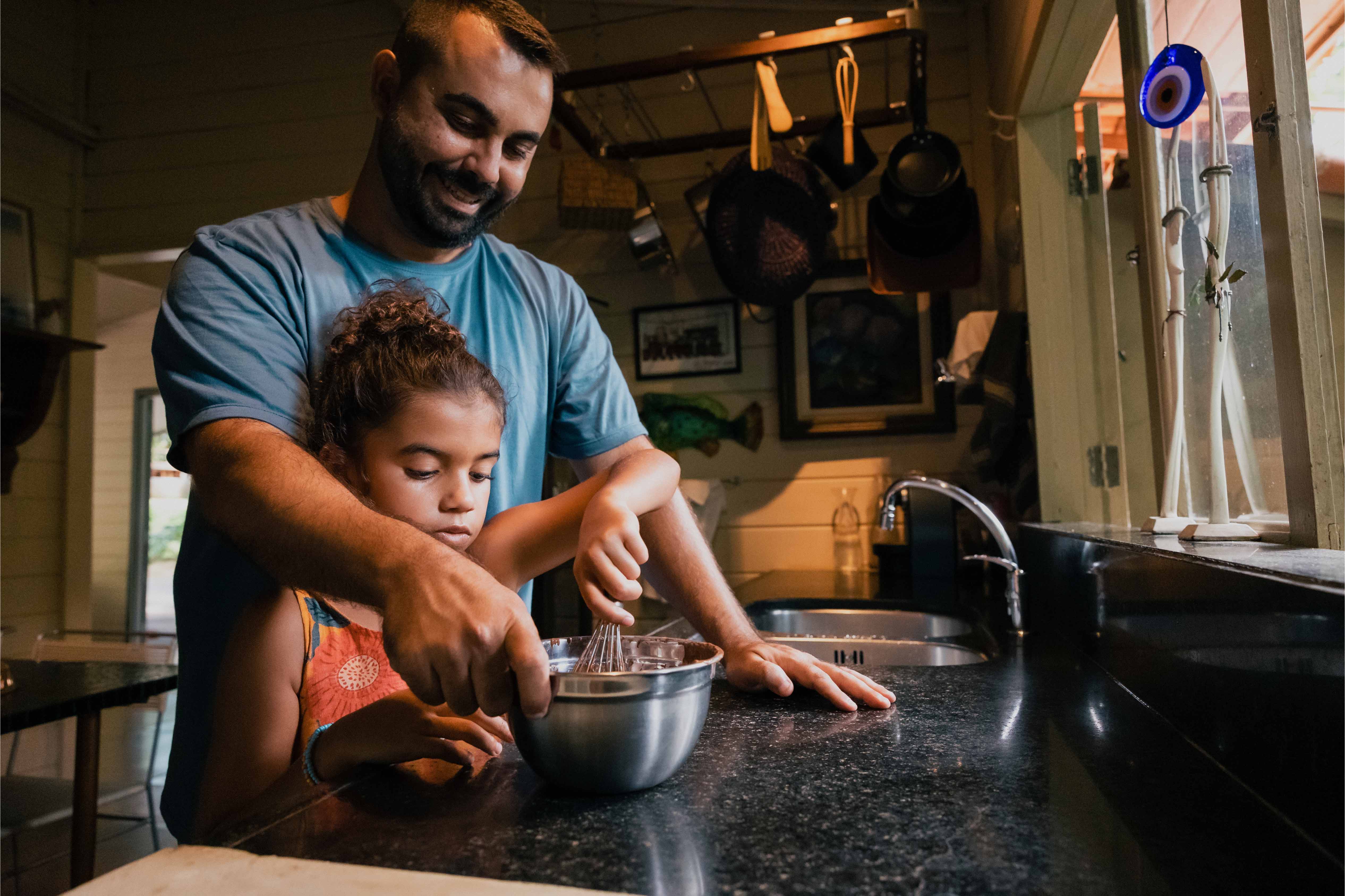 Smiling father helping his daughter whisk ingredients in a mixing bowl inside a warmly lit kitchen