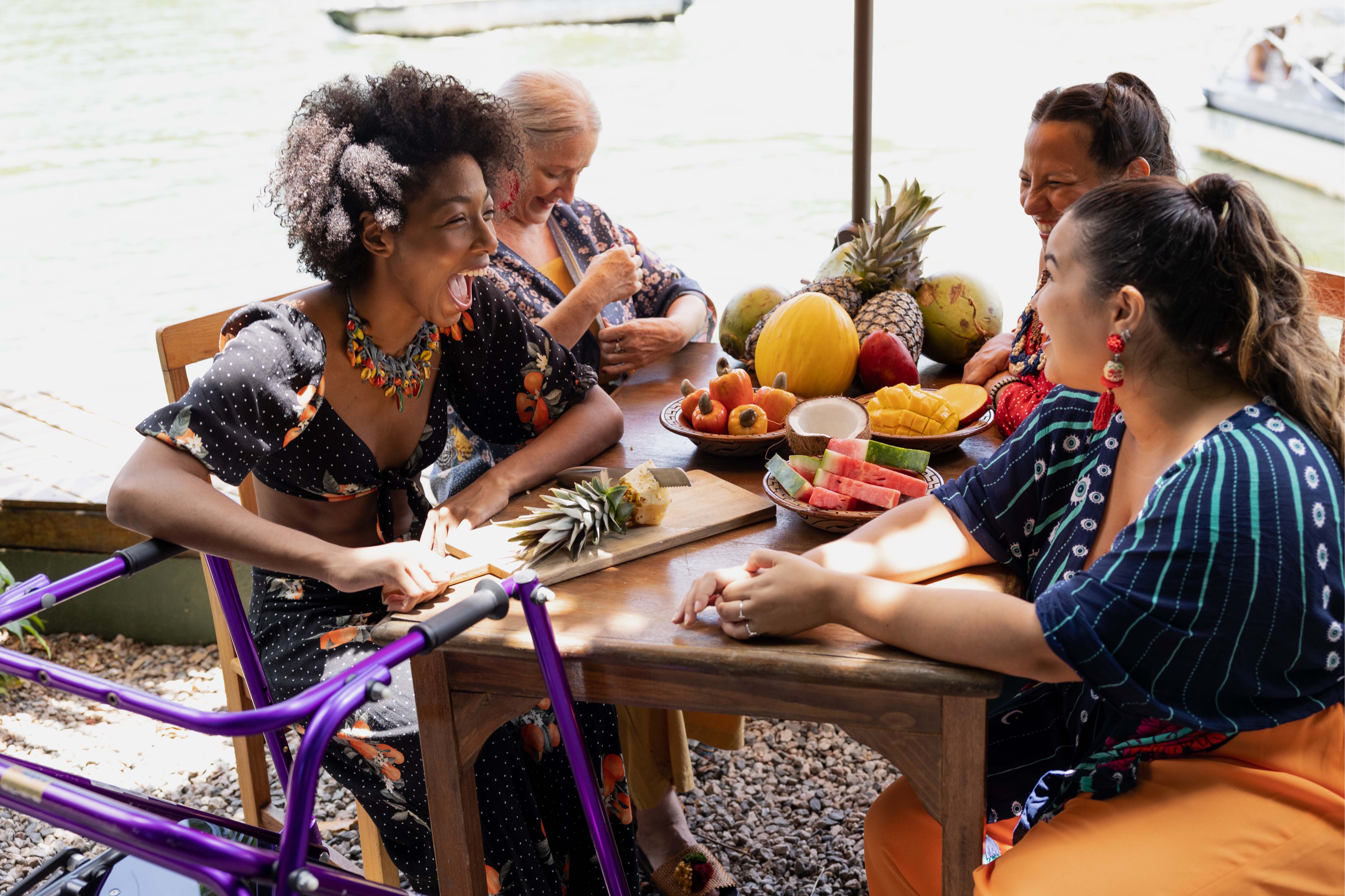 Diverse group of women laughing and sharing tropical fruit at an outdoor table near the water