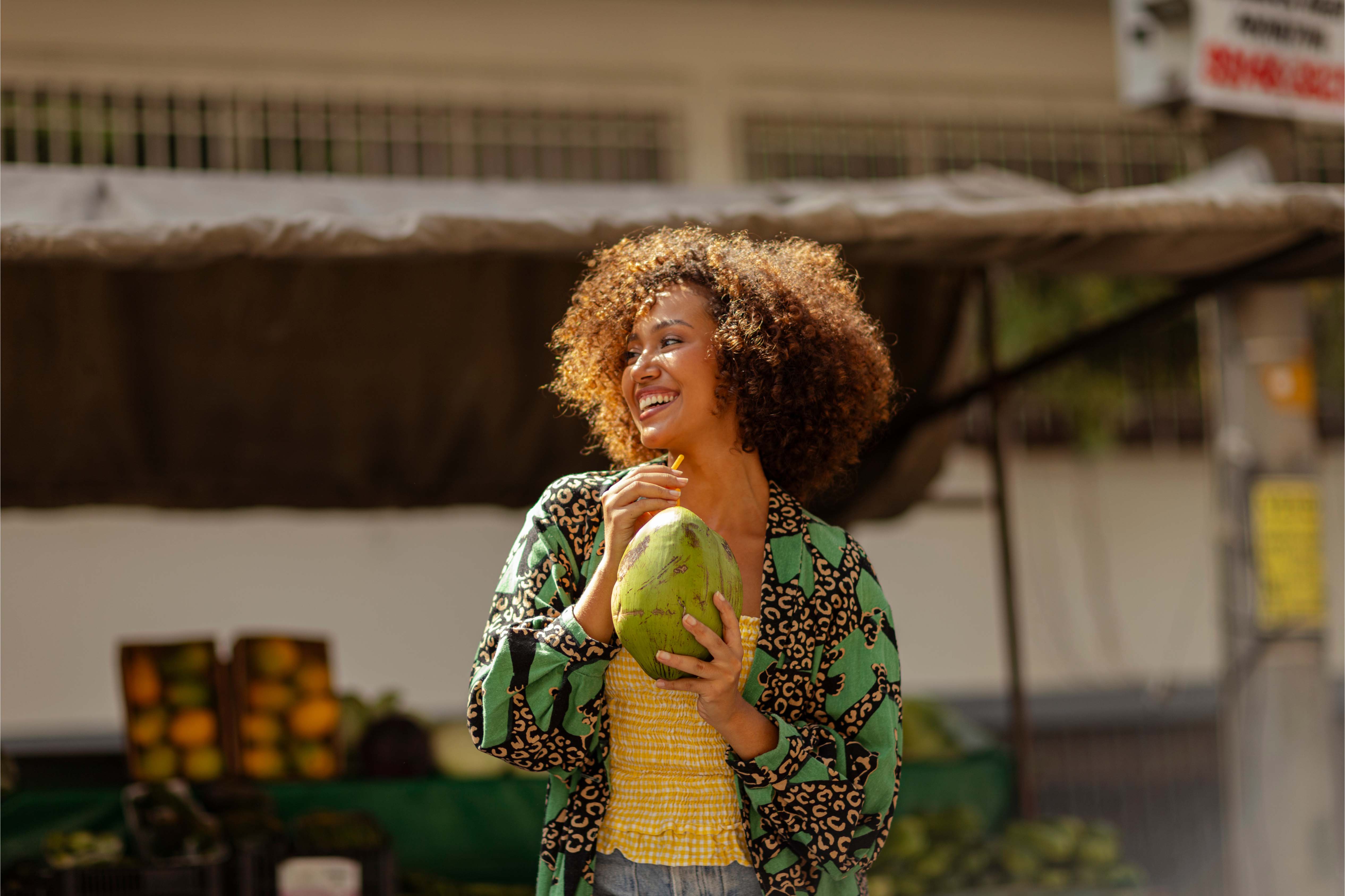 Happy woman holding a coconut with a straw while standing at an outdoor fruit market