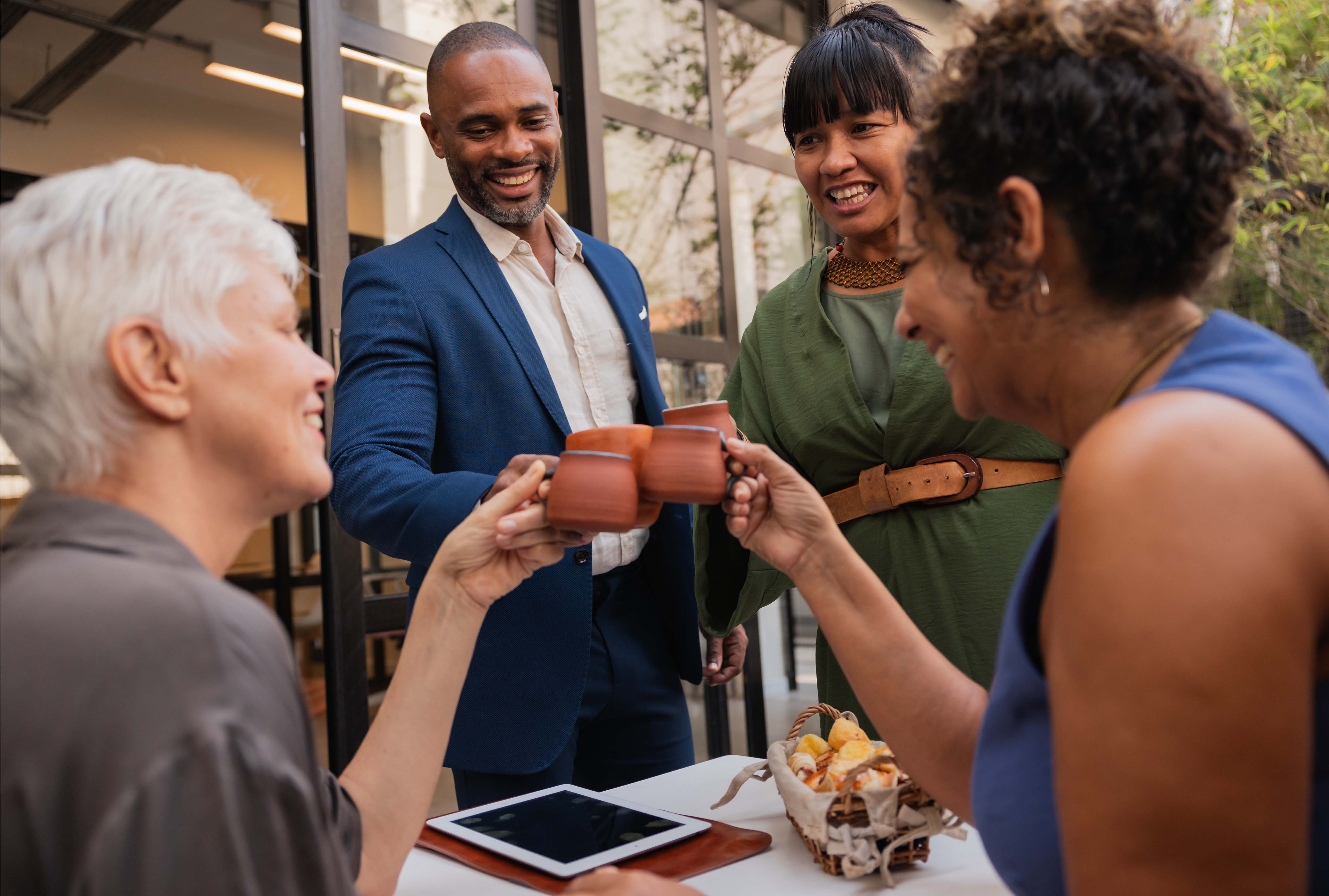Smiling colleagues raising mugs in a toast around a table with snacks and a tablet during an informal team meeting