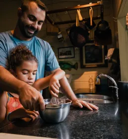 Smiling father helping his daughter whisk ingredients in a mixing bowl inside a warmly lit kitchen