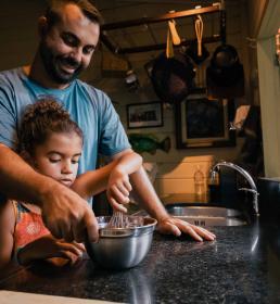 Smiling father helping his daughter whisk ingredients in a mixing bowl inside a warmly lit kitchen