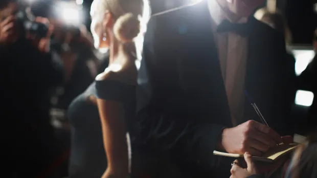 A man in a tuxedo signs an autograph book while a blonde woman in a ballgown is photographed by paparazzi behind him