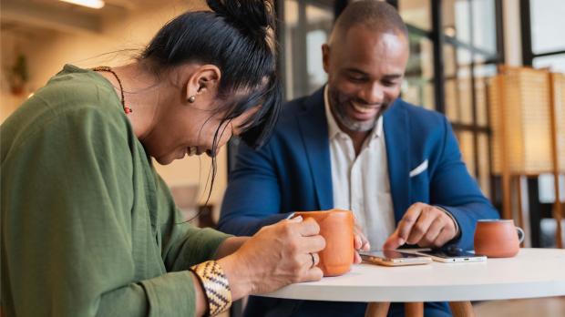 Man and woman smiling and laughing while having coffee and looking at a phone during a relaxed office catch-up