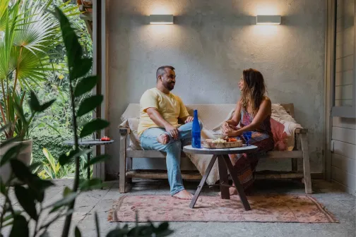Man and woman sitting on a bench outdoors, sharing food and conversation in a cosy, plant-filled veranda